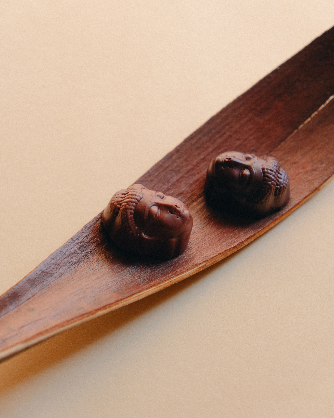 Dve praline u obliku Buddhe od mlečne i tamne čokolade položene na suvi list drveta, u toplom prirodnom osvetljenju.