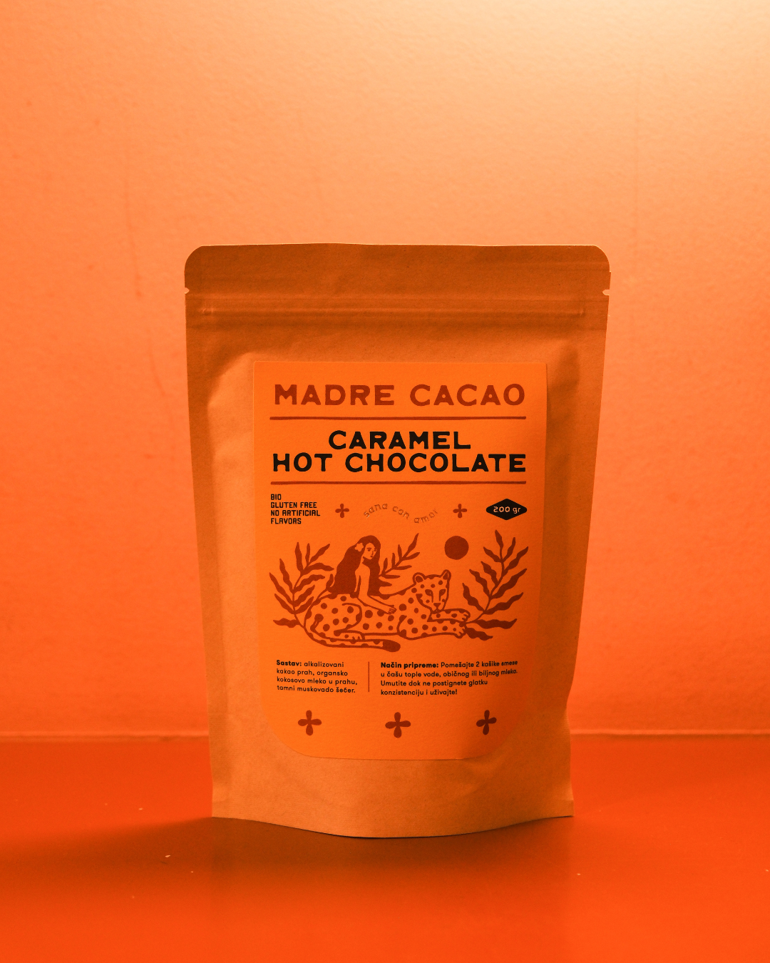 Paket Madre Cacao Caramel Hot Chocolate u kraft pakovanju sa žutom etiketom i ilustracijom žene i jaguara, fotografisan na toploj narandžastoj pozadini.