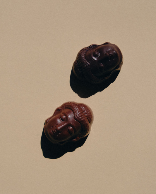Dve čokoladne praline u obliku Buddhe, napravljene od mlečne i tamne čokolade, na bež pozadini pod blagim svetlom.