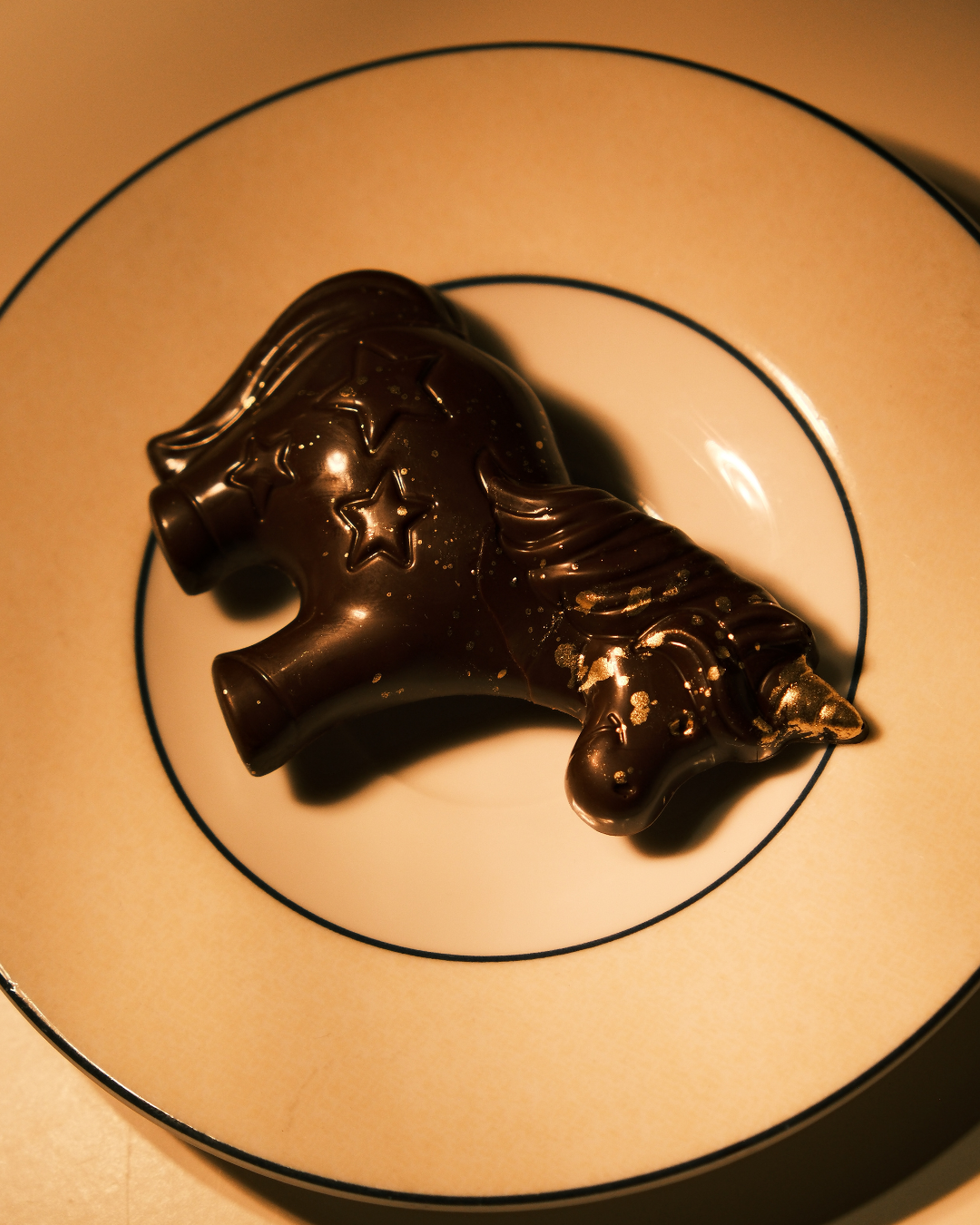 Pralina u obliku jednoroga od tamne čokolade sa zlatnim detaljima, položena na elegantan porcelanski tanjir sa tamnim obodom.