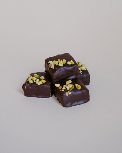 Praline od crne čokolade sa tahini pastom i pistaćima – gomilica kockica, posuto seckanim pistaćima, na svetloj pozadini