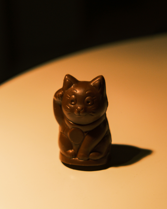 Lucky Cat pralina od mlečne čokolade na toplom svetlu – figura mačke koja maše, glazura sjajna.