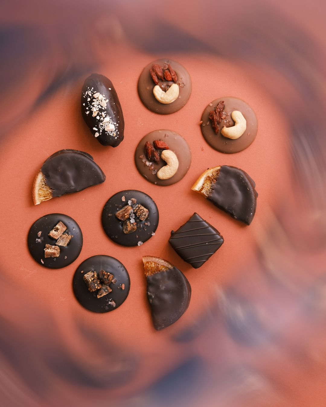 Krupni plan ručno pravljenih veganskih pralina od crne čokolade sa sušenim voćem, orašastim plodovima i kandiranom pomorandžom, raspoređenih na toploj smeđoj podlozi.