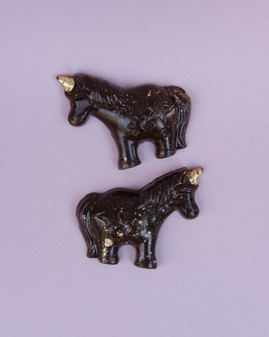 Dve praline u obliku jednoroga od crne čokolade sa zlatnim detaljima, položene na svetloljubičastoj pozadini.