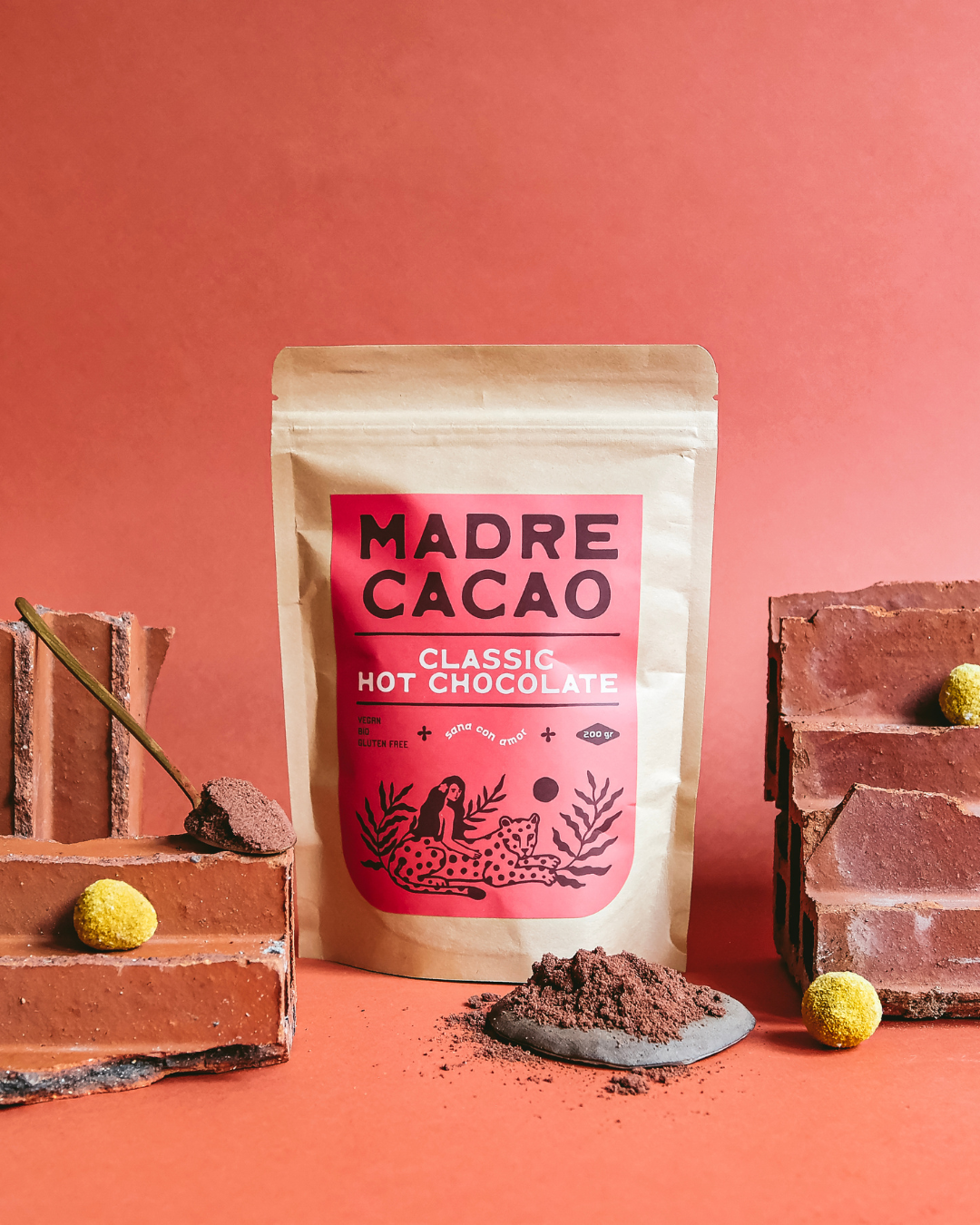 Paket Madre Cacao Classic Hot Chocolate na crvenoj pozadini, okružen kockama čokolade i prahom kakaoa — prirodna, veganska topla čokolada bez glutena.