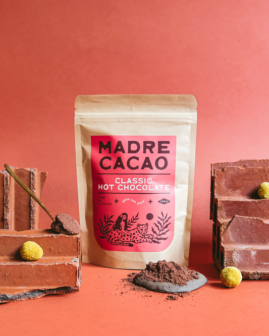 Paket Madre Cacao Classic Hot Chocolate na crvenoj pozadini, okružen kockama čokolade i prahom kakaoa — prirodna, veganska topla čokolada bez glutena.