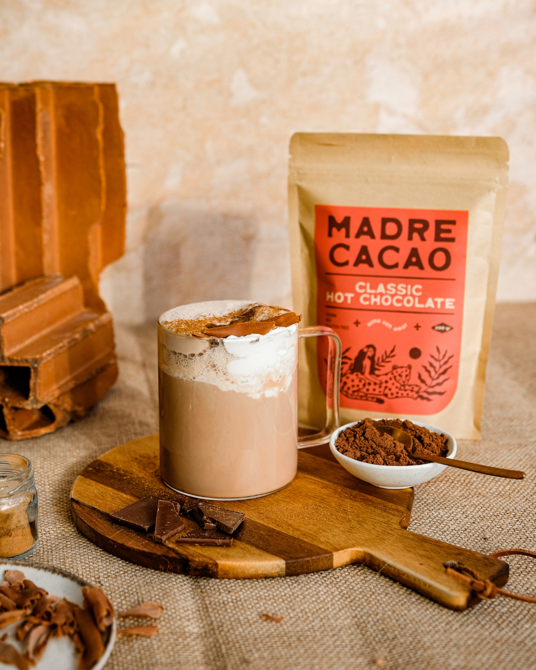 Šolja kremaste tople čokolade pored pakovanja Madre Cacao Classic Hot Chocolate, sa činijom kakao praha i komadićima čokolade na drvenoj dasci.