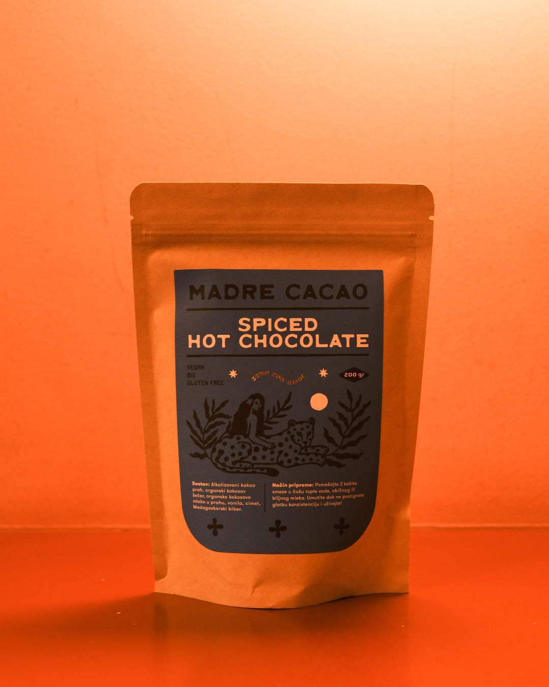 Paket Madre Cacao Spiced Hot Chocolate u kraft pakovanju sa tamnoplavom etiketom i ilustracijom žene i jaguara, fotografisan na toploj narandžastoj pozadini.