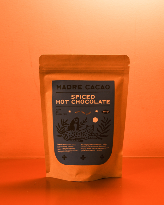 Paket Madre Cacao Spiced Hot Chocolate u kraft pakovanju sa tamnoplavom etiketom i ilustracijom žene i jaguara, fotografisan na toploj narandžastoj pozadini.